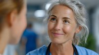 Mature Caucasian female healthcare professional with gray hair smiling warmly during conversation with colleague in modern medical facility environment.