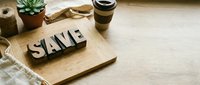 Wooden letters spelling SAVE on cutting board with coffee cup and succulent plant on natural wood table surface for financial planning concepts.