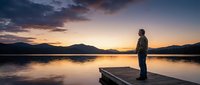 Man standing on wooden dock overlooking calm lake at sunset with mountain silhouettes and dramatic sky reflecting in water for meditation and solitude concepts.