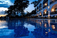 Luxurious resort swimming pool at dusk with illuminated Mediterranean-style building facade, reflecting lights and dramatic blue hour sky with clouds.