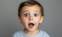 Surprised young child with wide open blue eyes and mouth expressing amazement and wonder against gray background, wearing light sweater. For emotional concepts.