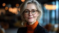 Confident mature businesswoman with blonde hair and glasses wearing orange turtleneck and dark blazer against blurred bokeh background. Professional corporate portrait.
