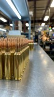 Rows of brass ammunition cartridges with copper bullets lined up on metal surface in manufacturing facility with industrial lighting and blurred background.