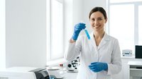 Young female scientist of Caucasian ethnicity wearing white lab coat and blue latex gloves, holding test tube in modern laboratory setting with microscope and equipment, suitable for research, healthcare, and education content.