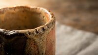 Rustic ceramic pottery bowl with weathered texture and earthy tones on wooden surface, handcrafted artisan craftsmanship for home decor and kitchen use.