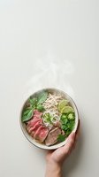 Overhead view of steaming bowl of Asian noodle soup with sliced beef, fresh herbs, lime, and rice noodles held by hand against neutral background.