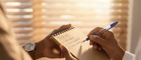 Professional businessman writing notes in spiral notebook with pen during morning sunlight through window blinds for business planning and productivity.