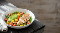 Grilled chicken breast sliced over fresh vegetables and greens in white bowl on wooden surface for healthy eating and nutrition content.