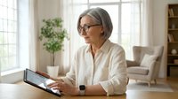 Mature businesswoman with gray hair and glasses using digital tablet at home office desk, professional remote work and technology concept.