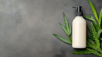 Blank beige cosmetic pump bottle with green aloe vera leaves on dark gray concrete background. Mockup for natural skincare products and organic beauty treatments.