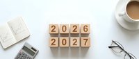 Wooden blocks showing 2026 and 2027 years on white desk with coffee cup, calculator, notebook and glasses for business planning and goal setting.