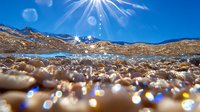 Underwater split view of sunlit ocean waves with sparkling bokeh reflections on sandy bottom against bright blue sky and sunburst through crystal clear water.