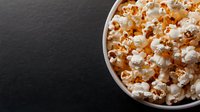 Fresh popcorn in white ceramic bowl against dark background, dramatic side lighting creates contrast between kernels and shadows, ready for movie night snack.