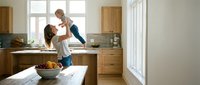 Happy mother lifting laughing toddler daughter in bright modern kitchen with wooden cabinets during playful morning routine at home.