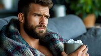 Young bearded Caucasian man relaxing on couch with hot beverage and marshmallows, wrapped in plaid blanket, expressing contemplative mood at home.