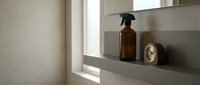 Amber glass spray bottle and round mirror on gray floating shelf near window with natural light and neutral wall decor.