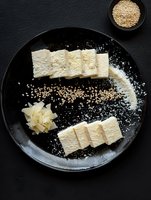 Sliced white marshmallow treats dusted with powdered sugar and sesame seeds on black ceramic plate, served with ginger garnish for Asian dessert presentation.