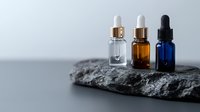 Essential oil dropper bottles in clear, amber and blue glass arranged on dark stone surface against neutral background, minimalist spa composition.