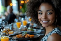 Young African American woman with natural curly hair smiling at restaurant table with orange juice and elegant place settings during brunch or dinner gathering.