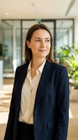 Professional woman with long brown hair and Caucasian ethnicity wearing navy blazer and white shirt in modern office lobby with glass partitions and plants, suitable for corporate, business, and professional portraits.
