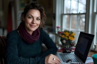 Woman with dark wavy hair and olive complexion wearing burgundy scarf and teal sweater working on laptop at home office desk by window.