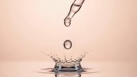 Water droplet creating splash crown with falling drops against soft pink background capturing motion and fluid dynamics in macro photography.