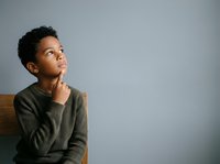 Young boy with dark curly hair and medium skin tone wearing olive green sweater, sitting thoughtfully with hand to chin, gazing upward against neutral gray background for educational and lifestyle content.