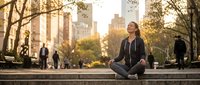 Young woman meditating in lotus position on city steps during golden hour sunset with urban skyline and people walking in background for wellness content.