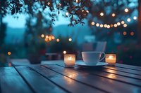 White coffee cup and glowing candles on wooden table at dusk, with bokeh fairy lights and foliage creating cozy outdoor atmosphere. Ideal for lifestyle content.