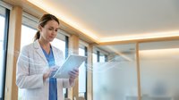 Professional female doctor in white coat reviewing medical documents in modern hospital corridor with natural lighting and glass walls.