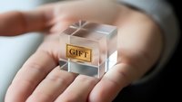 Transparent glass cube with golden gift label floating above open palm, conceptual present or reward symbol for business and celebration designs.