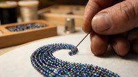 Artisan hand-stitching iridescent blue beads onto white fabric in embroidery hoop, creating elegant curved pattern. Craft and textile art concept.