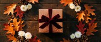 Autumn gift box with burgundy ribbon surrounded by orange maple leaves, white flowers and pine cones on rustic wooden background for seasonal celebrations.