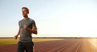 Athletic man with reddish-blonde hair jogging on outdoor track at sunset, wearing gray shirt. Fitness, training, and sports concept for wellness and healthy lifestyle content.