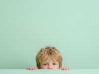 Curious young boy with blonde hair peeking over white surface against mint green background, expressing wonder and playfulness. Suitable for parenting, education, and childcare marketing materials.