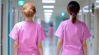 Two young female healthcare workers wearing pink scrubs walking down a modern hospital corridor with fluorescent lighting, viewed from behind.