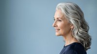 Mature woman with long gray hair and fair complexion, wearing dark clothing, smiling in profile against neutral blue background. Suitable for healthcare, wellness, aging, lifestyle and corporate use.