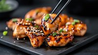 Glazed teriyaki chicken wings garnished with sesame seeds and green onions on black plate, close up view with chopsticks picking up a piece, dark moody background.