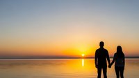 Romantic couple silhouettes holding hands watching golden sunset over calm water creating peaceful moment together for love and relationship concepts.