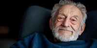 Smiling elderly Caucasian man with white beard and blue sweater against dark background shows genuine happiness and contentment in candid portrait.
