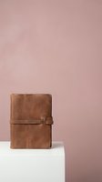Vintage brown leather journal notebook with strap closure on white pedestal against soft pink background for stationery and writing concepts.