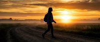 Silhouette of a young adult male jogging on a rural dirt road during golden hour sunrise or sunset, wearing athletic gear and a backpack, with expansive countryside landscape and dramatic sky.