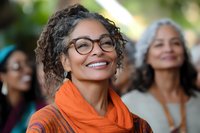 Mature African American woman with curly gray hair and glasses wearing bright orange scarf smiling outdoors, expressing joy and confidence. Group of diverse women in background.