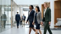 Three diverse professional businesswomen walking confidently through modern corporate office lobby, representing teamwork and leadership in workplace.