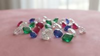 Collection of loose precious gemstones including emeralds, rubies, sapphires and diamonds scattered on pink surface for jewelry making and luxury design.