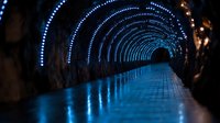 Illuminated tunnel with blue LED lights creating magical perspective effect. Curved stone pathway with reflective wet surface leads into dark distance.