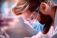 Bearded medical professional in protective face mask working in laboratory setting with dramatic lighting and soft pink-blue tones creating atmospheric mood.