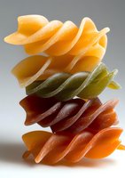 Colorful spiral pasta noodles stacked vertically showing yellow, green, red and orange varieties against gray background, dramatic lighting highlights texture.