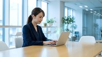 Young Asian businesswoman working on laptop in modern office conference room with natural light and glass windows for corporate presentations.