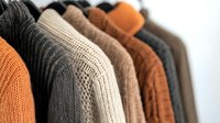 Cozy knitted sweaters in autumn colors hanging on rack against white wall. Warm wool and cotton pullovers arranged in row showing texture and patterns.